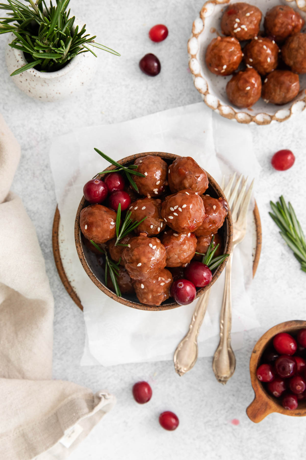 Overhead image of cranberry meatballs displayed in a bowl. Utensils are visible on the right side of the bowl, and herbs and cranberries are scattered around the dish. 