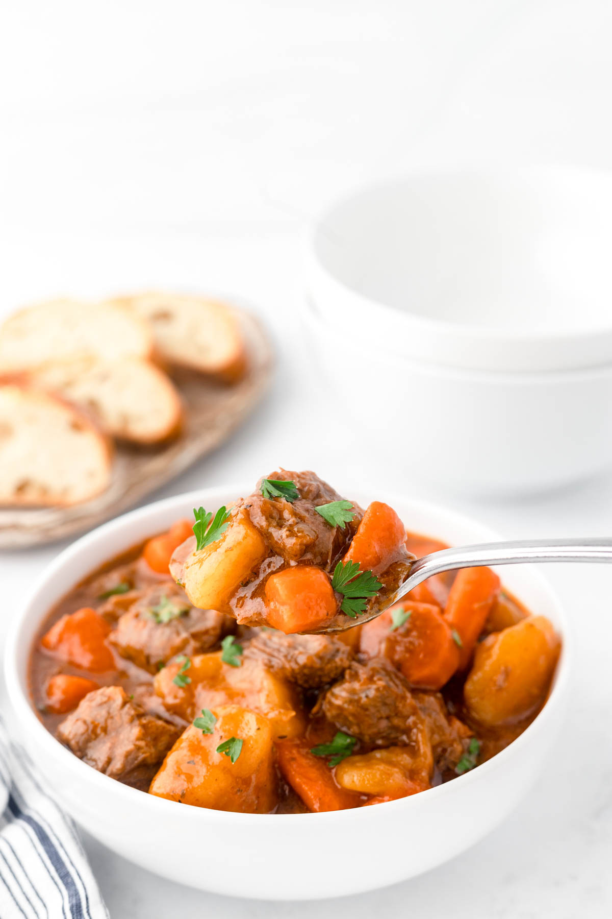 A bowl filled with beef stew is in the foreground, with a spoonful of stew raised closer to the camera. 