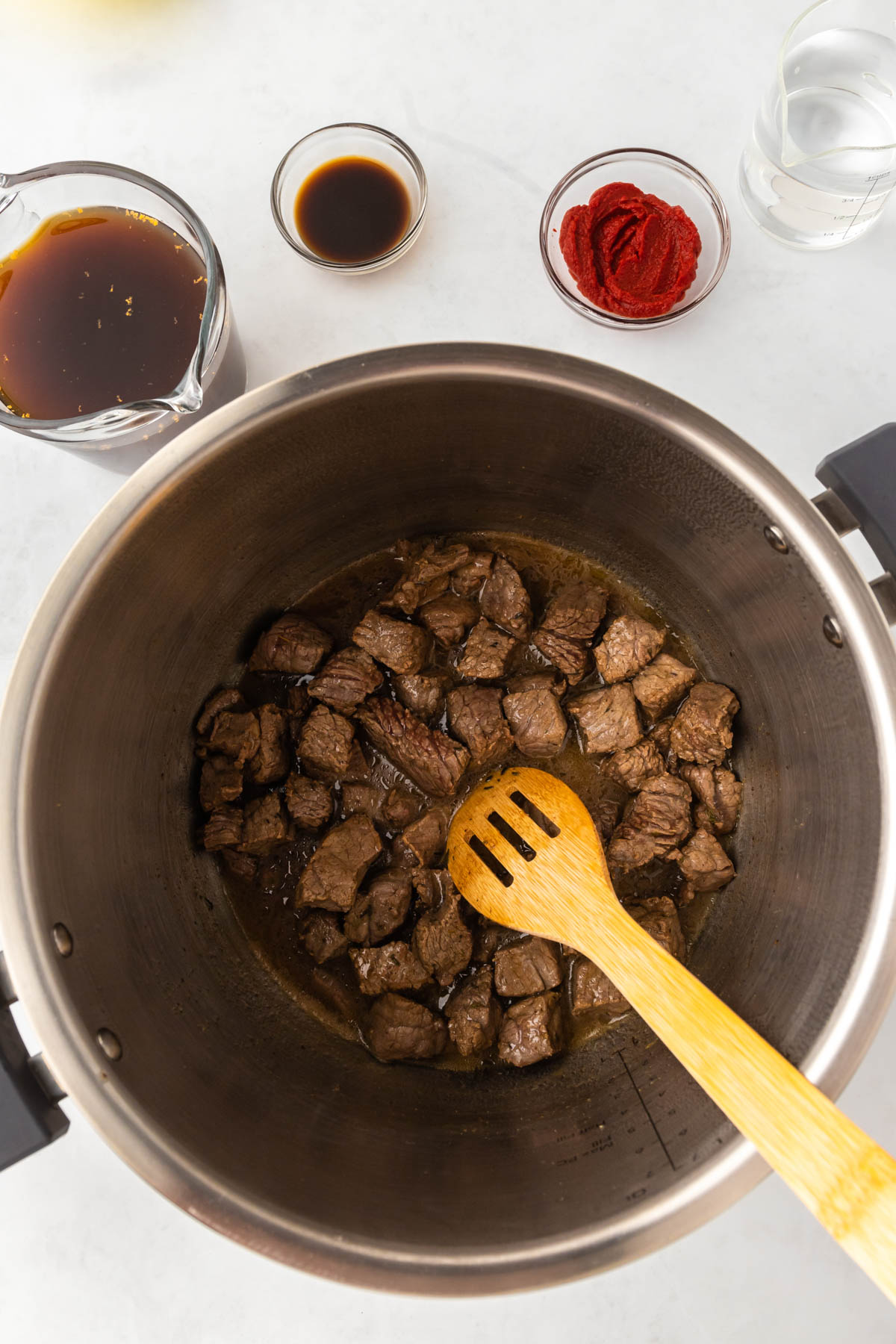 Beef is sauteed in an instant pot. A wooden spoon is visible in the pot, and spices in individual bowls are arranged around it.