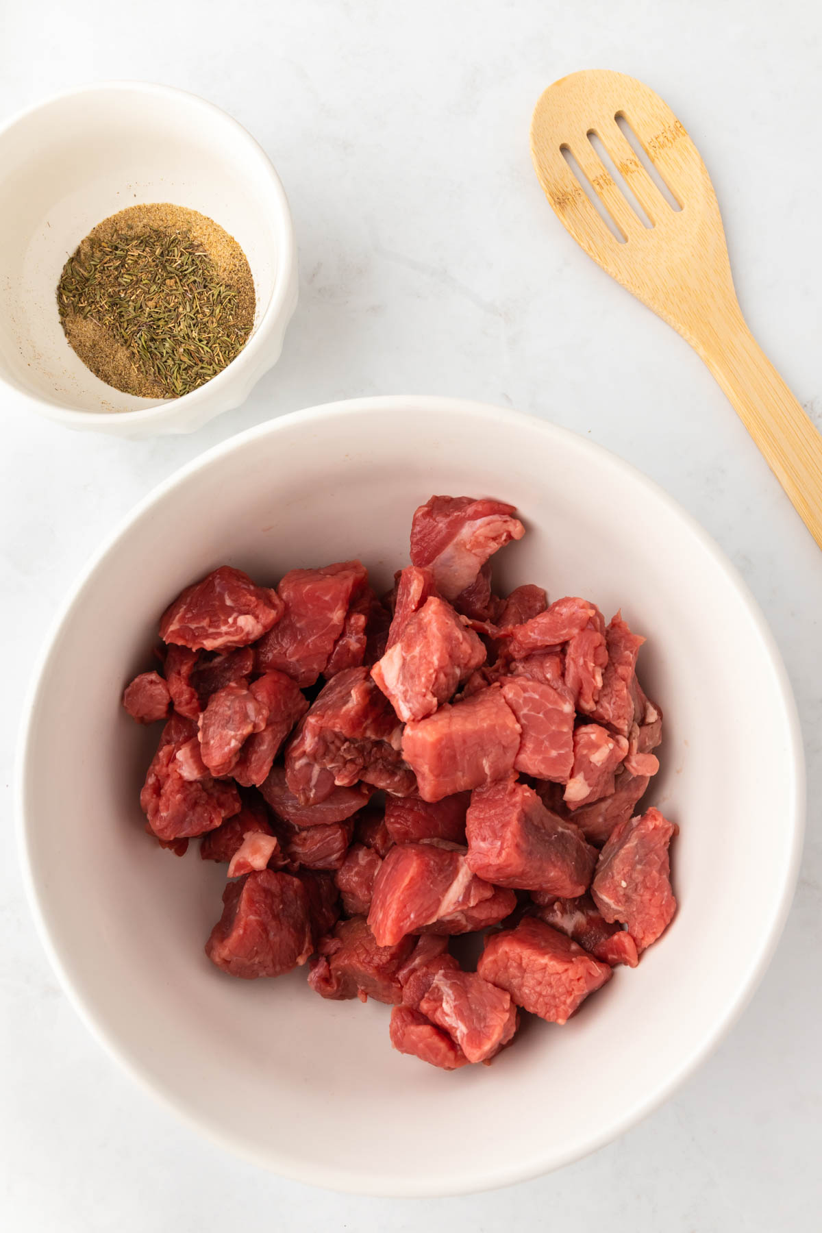 Cubed pieces of beef are in a bowl next to a small bowl with spices. 
