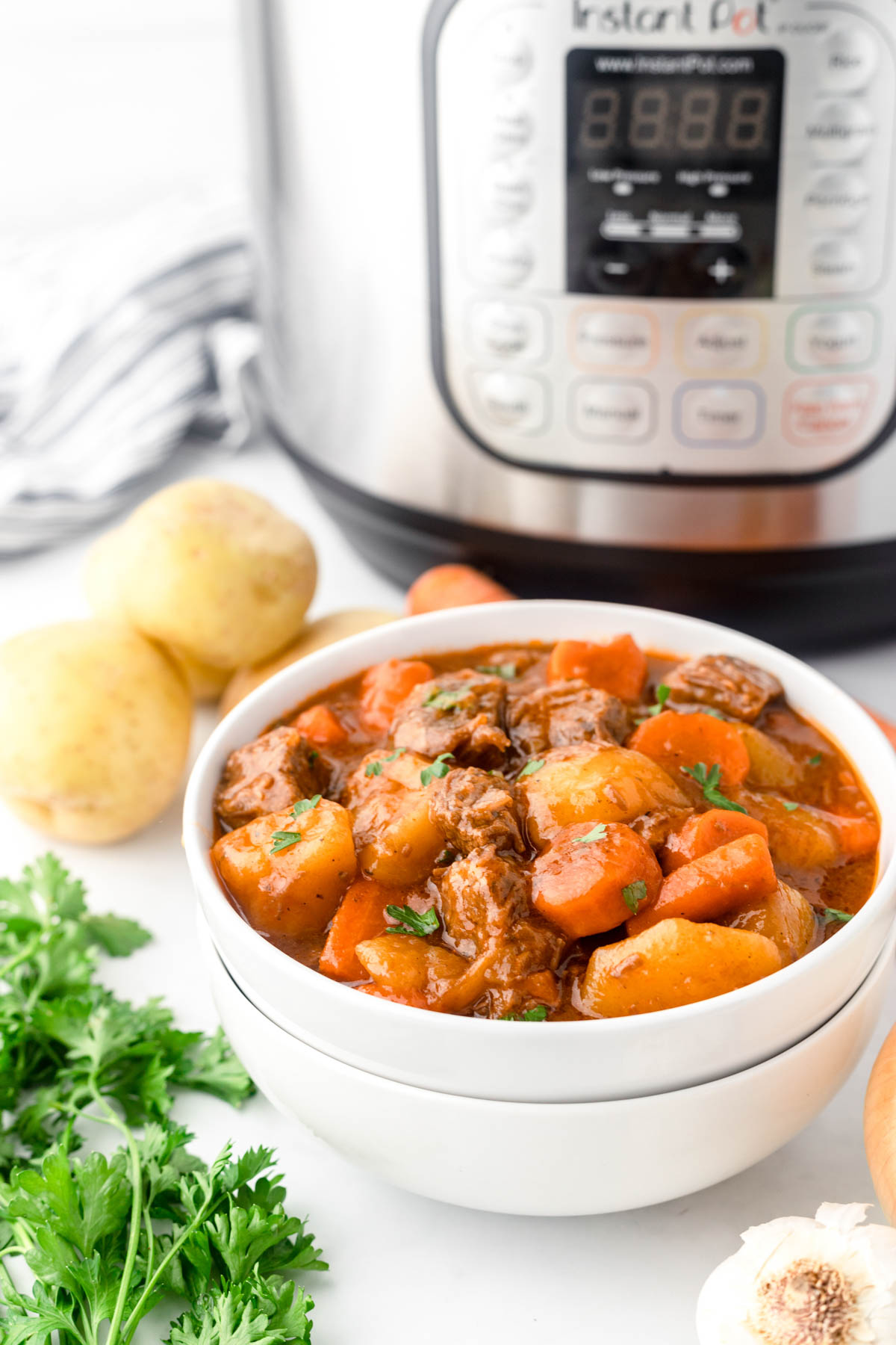 Cooked beef stew is displayed in a white bowl in the foreground. Behind it, an Instant Pot and vegetables are visible.