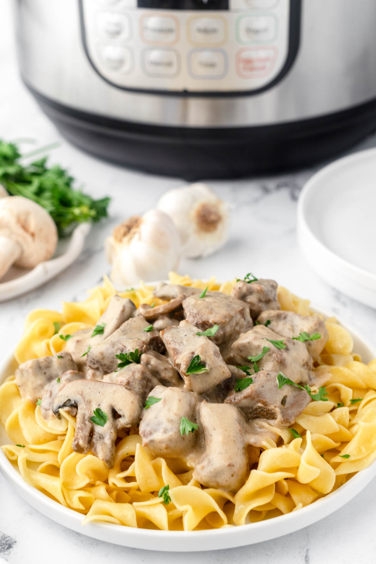 Beef chunks are coated in a stroganoff sauce and sprinkled with chopped parsley, served over cooked egg noodles. Mushrooms, garlic, herbs and an Instant Pot are visible in the background. 