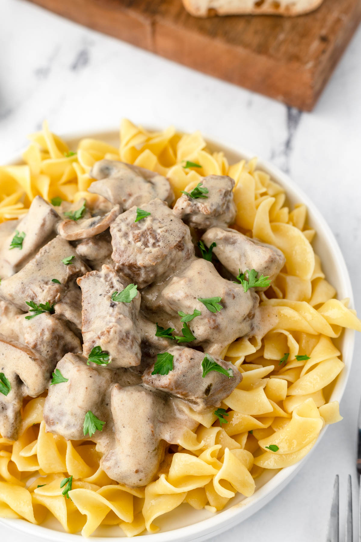 A dish of beef stroganoff is served over cooked egg noodles. The corner of a cutting board and sliced bread are visible in the top corner. 