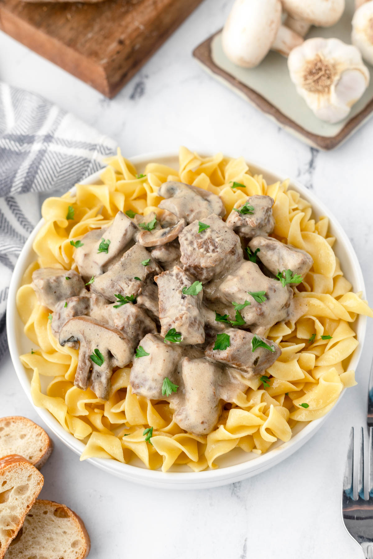 Overhead shot of a plate of egg noodles and beef stroganoff, with garlic cloves, sliced french bread and a dish cloth visible around the plate. 