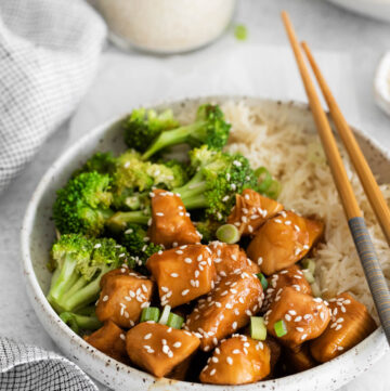 Close up of a bowl containing teriyaki chicken, broccoli and rice, with chopsticks displayed across the rim of the bowl.