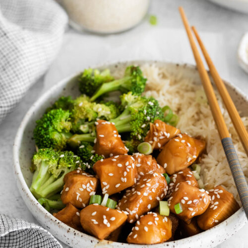 Close up of a bowl containing teriyaki chicken, broccoli and rice, with chopsticks displayed across the rim of the bowl.