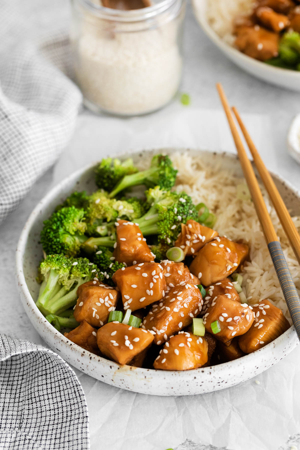 Close up of a bowl containing teriyaki chicken, broccoli and rice, with chopsticks displayed across the rim of the bowl. 