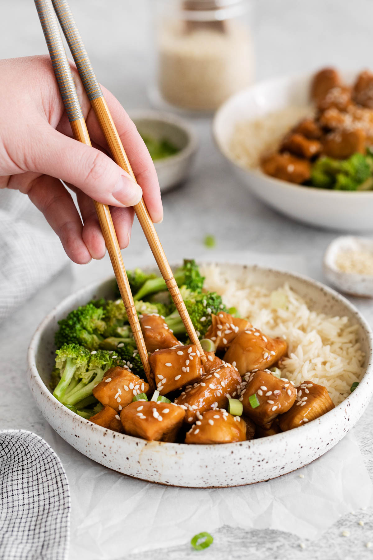 A bowl of teriyaki chicken, broccoli and rice is displayed in the foreground. Hands holding chopsticks are reaching for a piece of chicken. 