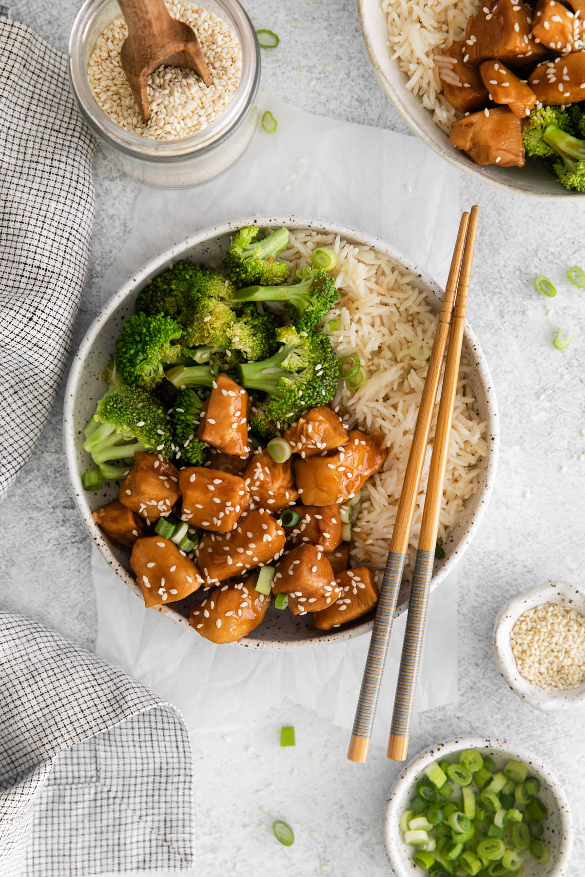 Overhead view of a bowl of teriyaki chicken, broccoli and rice with a pair of chopsticks resting on the side of the bowl. Small bowls filled with garnish are scattered around the main dish. 