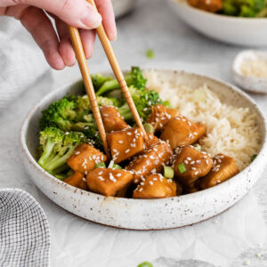 A plate of teriyaki chicken with a hand holding chopsticks to pick up a chunk of chicken.