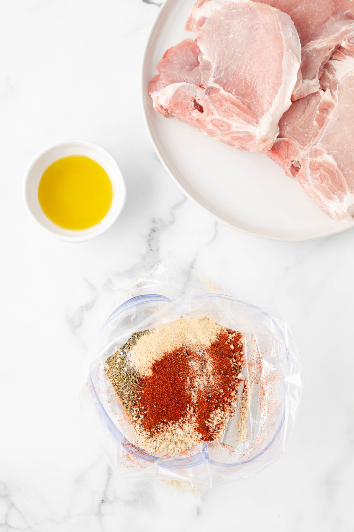 Spices are layered on top of a bag filled with panko breadcrumbs, displayed near a plate of uncooked pork chops and a small bowl of olive oil. 