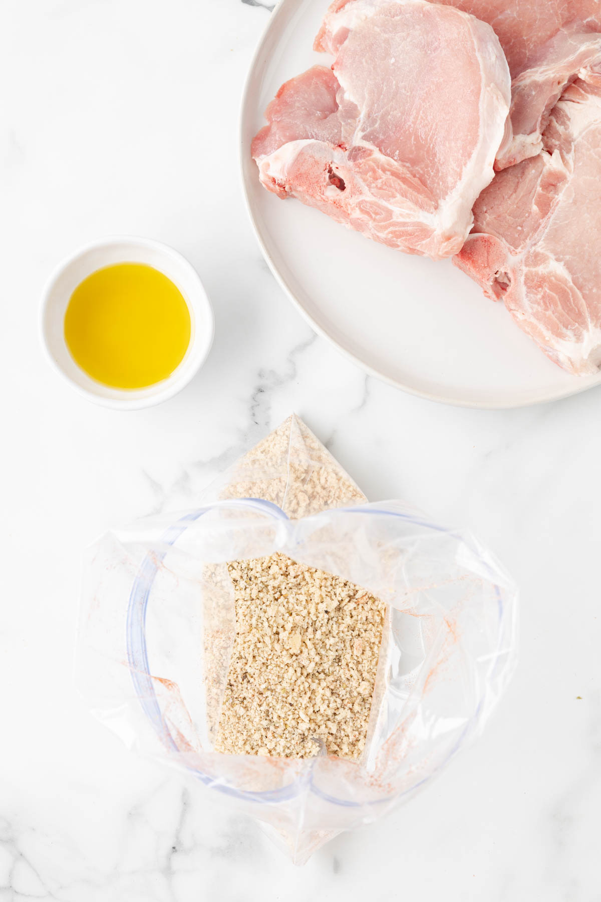A zip top bag is filled with breadcrumbs and is set on a surface. A plate of pork chops and a small bowl of olive oil are nearby. 