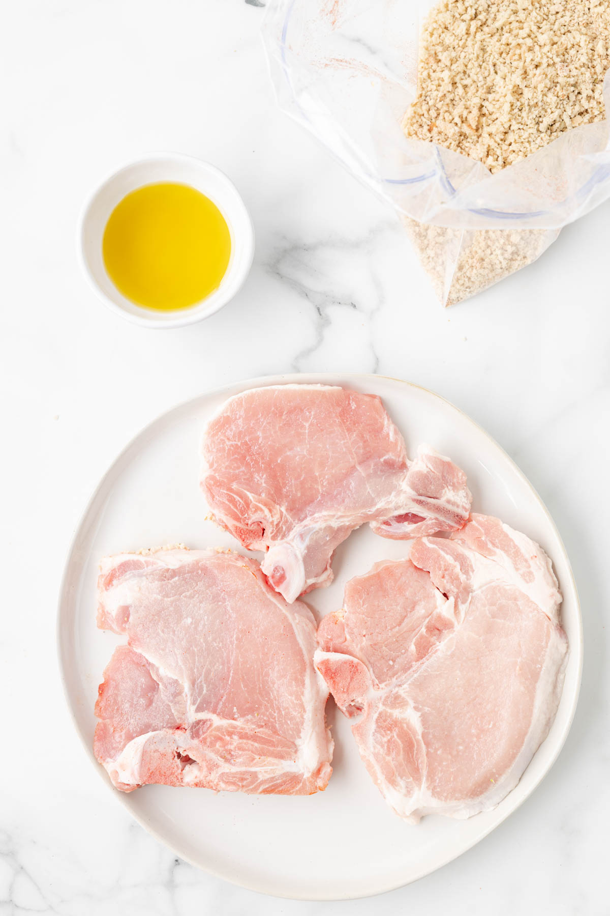 Pork chops are displayed on a white plate, with a small bowl of olive oil and a bag of bread crumbs and herbs in a zip top bag. 