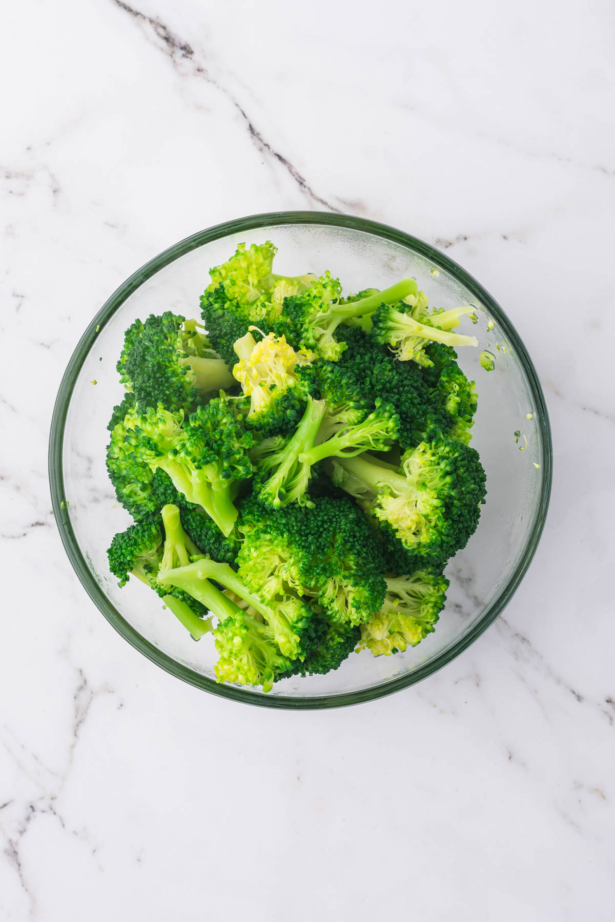 An overhead image of a clear glass bowl containing cooked broccoli florets. 