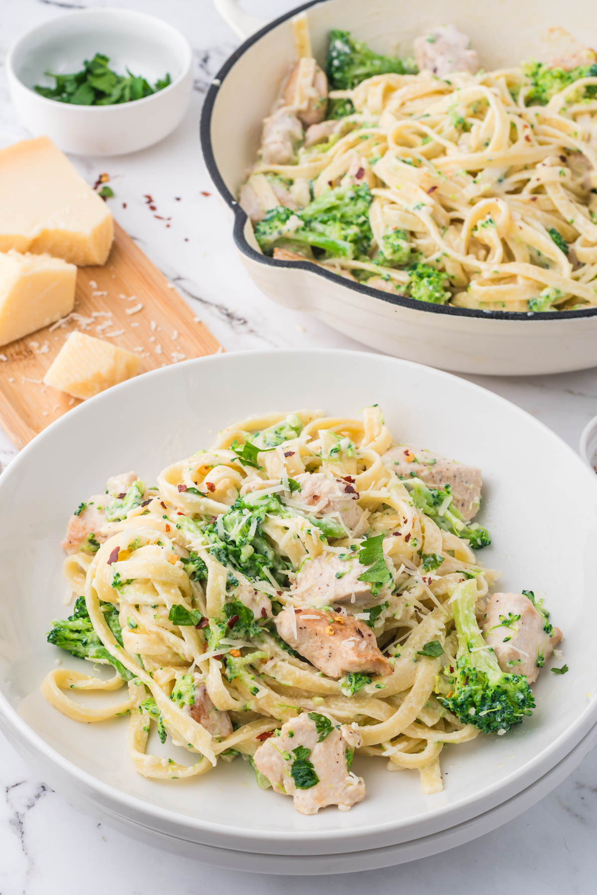 A closeup image of chicken and broccoli fettuccini alfredo on a white plate, with chunks of parmesan cheese, a bowl of herbs, and a pot containing more pasta in the background. 