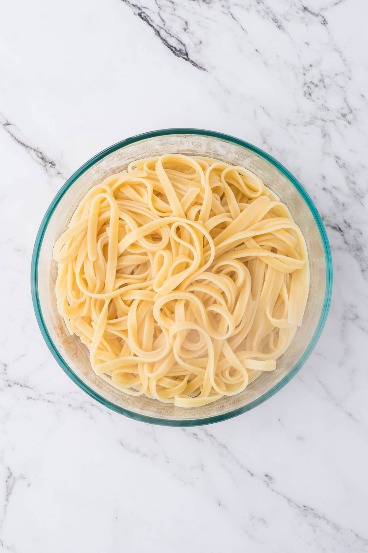 An overhead image of a clear glass bowl containing cooked pasta noodles. 