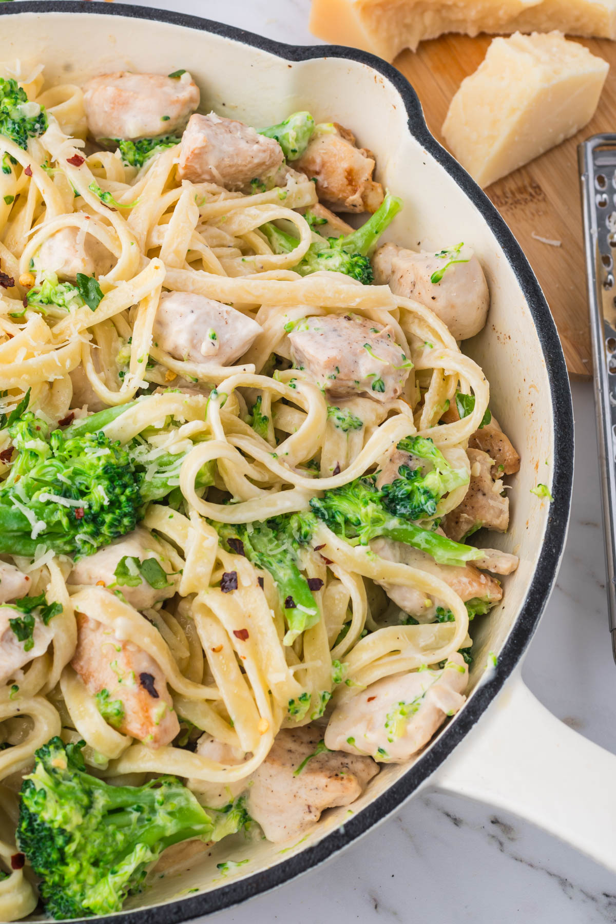 A closeup image of cooked chicken, noodles and broccoli in a creamy white sauce, in a pot. A metal grater and chunks of parmesan cheese are visible in the background. 