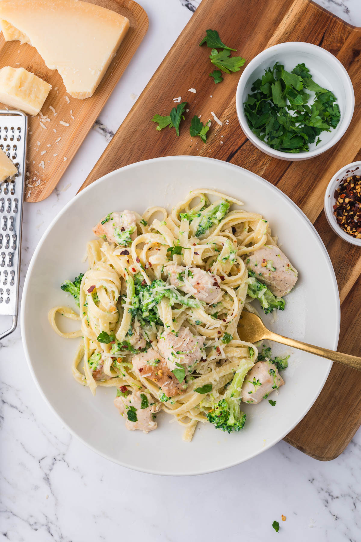 Overhead image of a plate of chicken fettuccini alfredo, with a cheese grater and bowls of herbs visible surrounding the plate, displayed on a wooden cutting board.