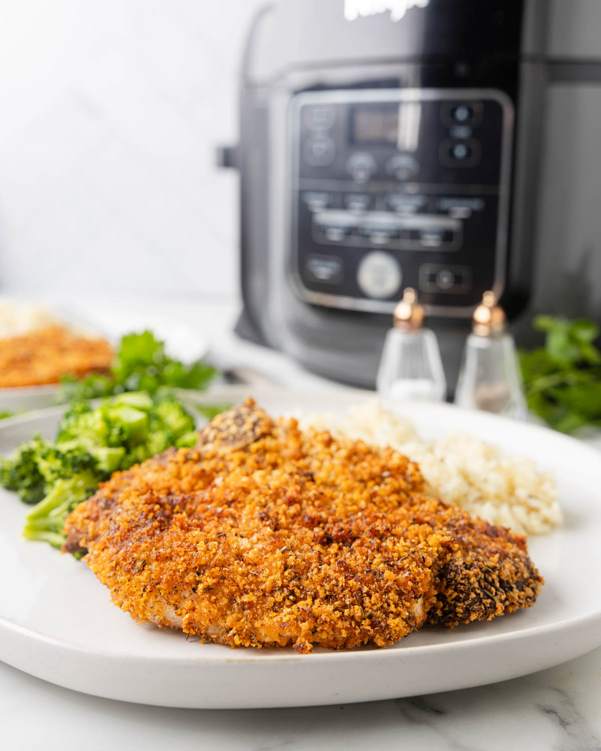 Side view of an air fryer fried pork chop on a serving plate with broccoli and rice. An air fryer is visible in the background. 