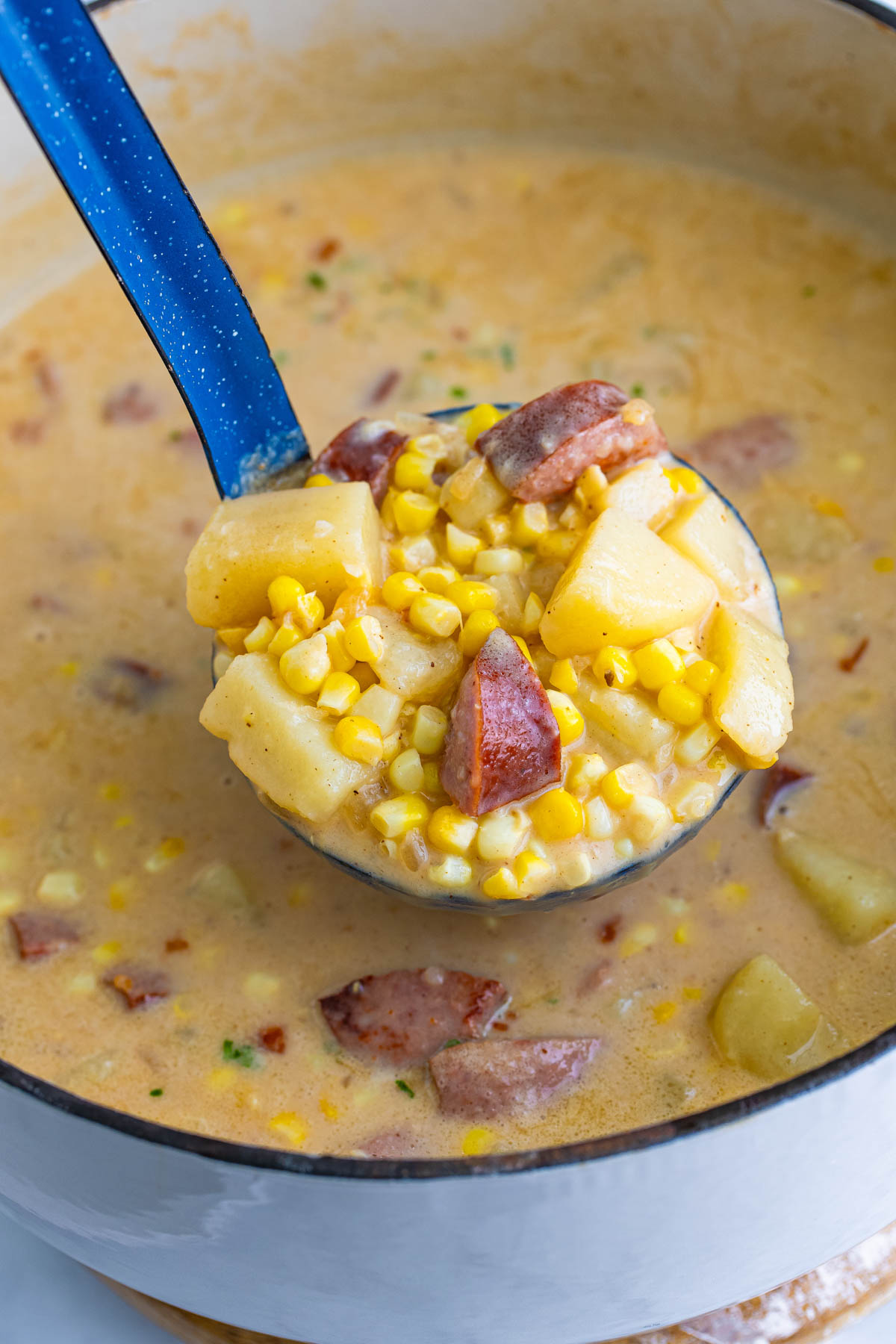 A large spoon is scooping sausage, potatoes and corn from a large stock pot.